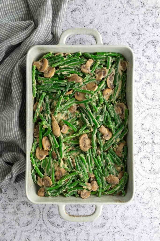 A rectangular baking dish filled with green bean casserole with sliced mushrooms and creamy sauce, placed on a light-colored surface next to a striped cloth.