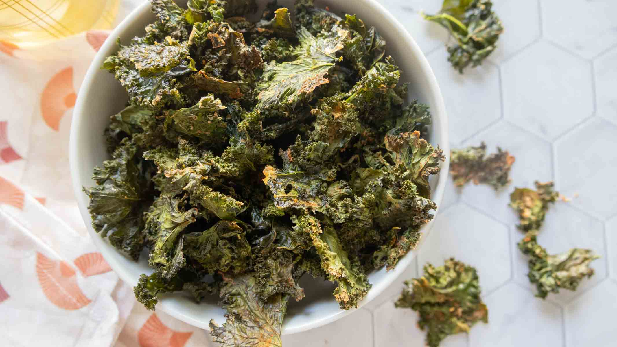 A white bowl filled with crispy baked kale chips sits on a white hexagon-tiled surface with a few chips scattered nearby.