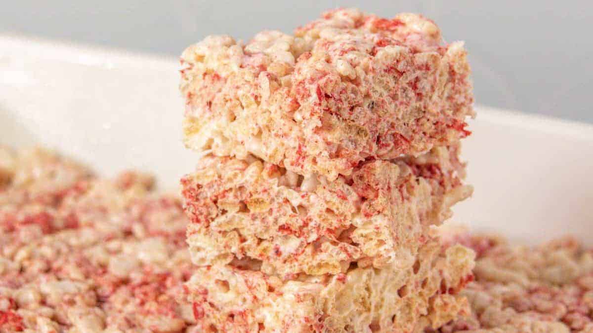 Three stacked Rice Krispies treats with visible red specks, likely strawberry-flavored, set against a blurred background of more treats.