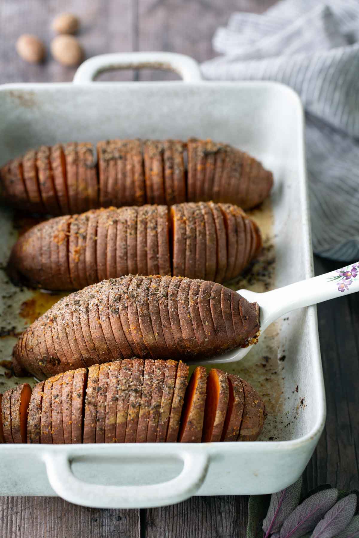 Four sliced, baked sweet potatoes with seasoning are arranged in a white rectangular baking dish on a wooden surface.