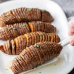 Four baked Hasselback sweet potatoes garnished with herbs on a white plate, with a spatula lifting one potato.