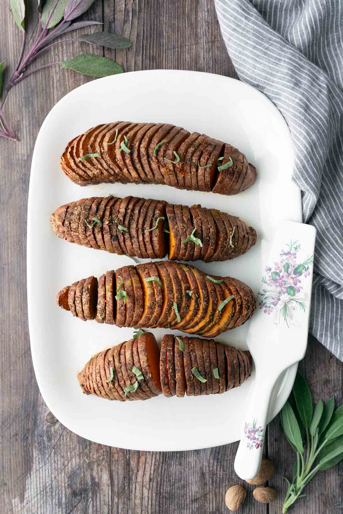 Four sliced, baked sweet potatoes garnished with herbs are arranged on a white platter with a floral-patterned server, set on a rustic wooden table.