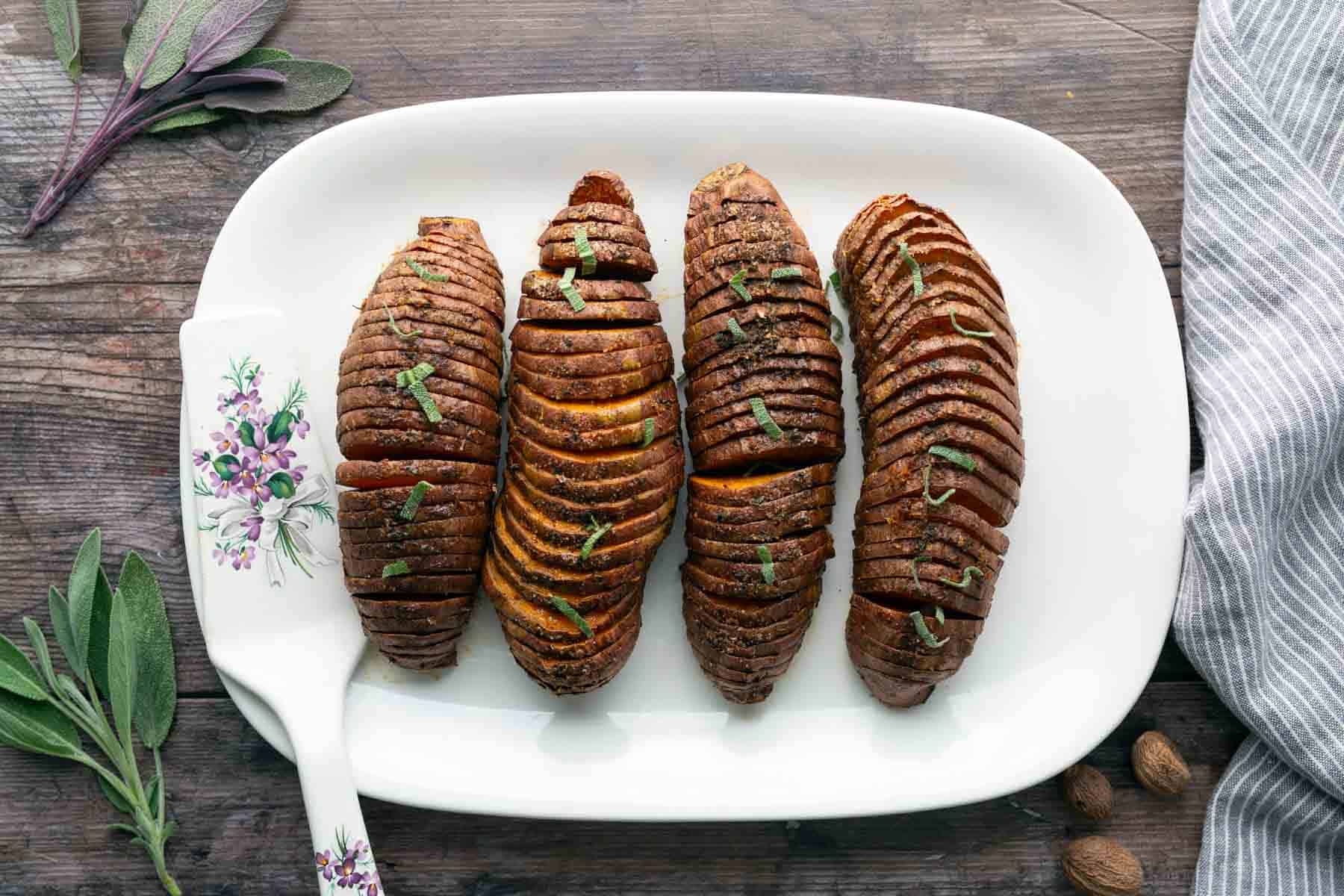 Four sliced, roasted sweet potatoes are arranged on a white platter, garnished with herbs, with a decorative serving spatula and fresh sage nearby.