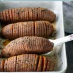 A baking dish with four sliced hasselback sweet potatoes topped with seasoning, one being served with a white spoon.