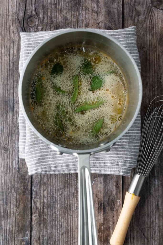 A saucepan with bubbling brown butter and sage leaves sits on a striped cloth, next to a metal whisk on a wooden surface.