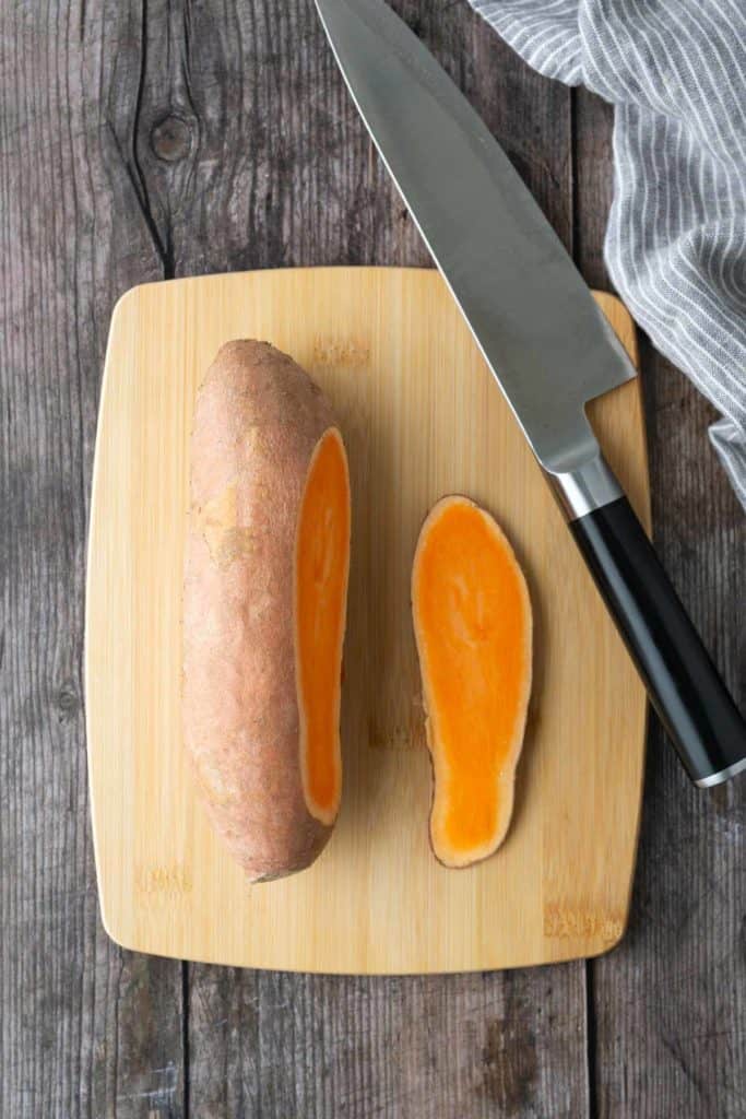 A halved sweet potato and a large knife on a wooden cutting board, set on a rustic wooden surface with a gray-striped cloth nearby.
