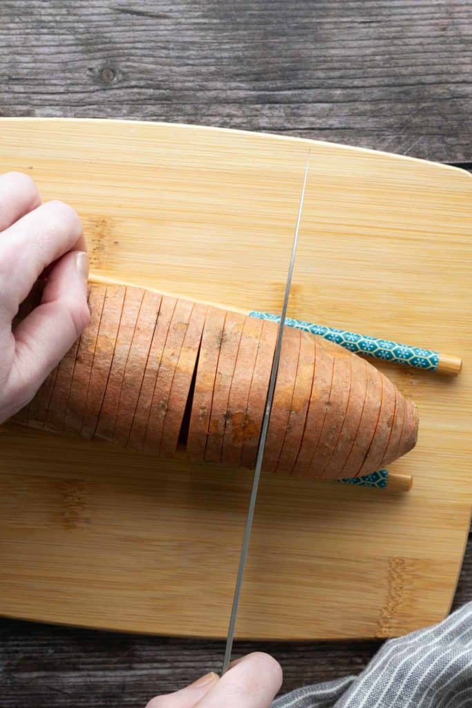 A hand slices a sweet potato on a wooden cutting board, using two chopsticks on either side to prevent cutting all the way through.