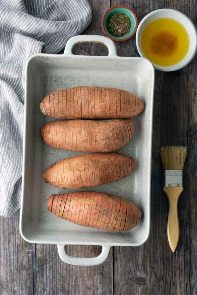 Four sliced sweet potatoes are arranged in a white baking dish. Nearby are a bowl of melted butter, a small bowl of herbs, a basting brush, and a gray striped towel.