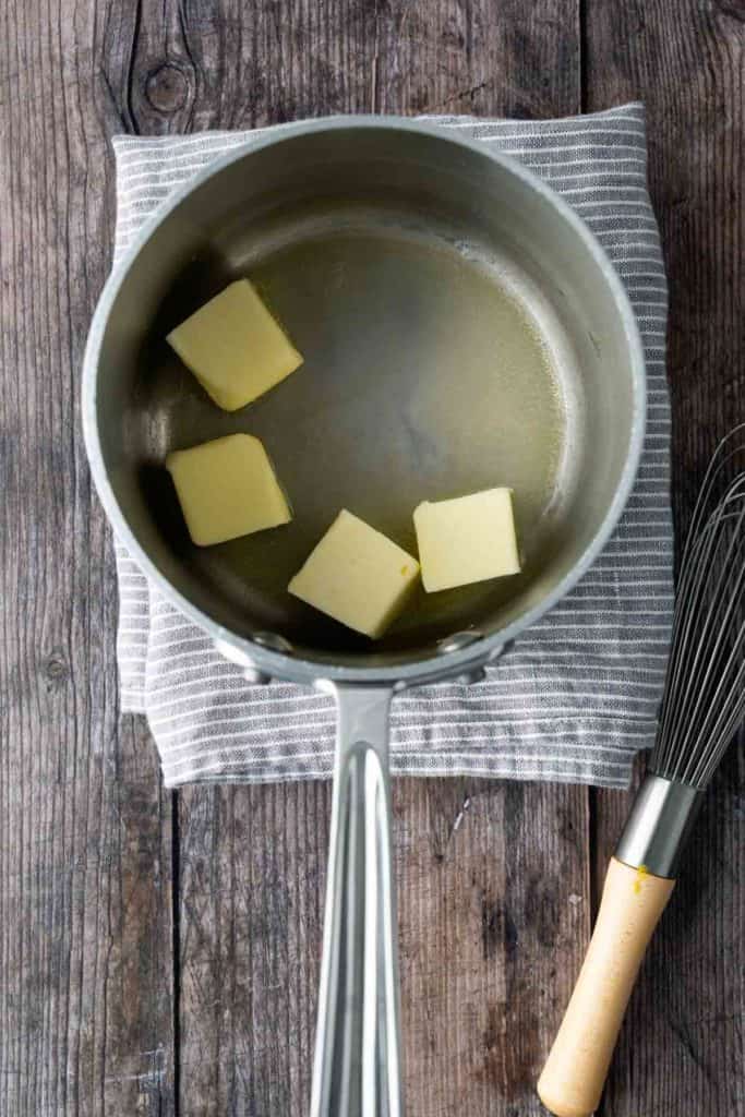 A saucepan with four cubes of butter partially melted, placed on a gray striped cloth on a wooden surface, next to a metal whisk with a wooden handle.