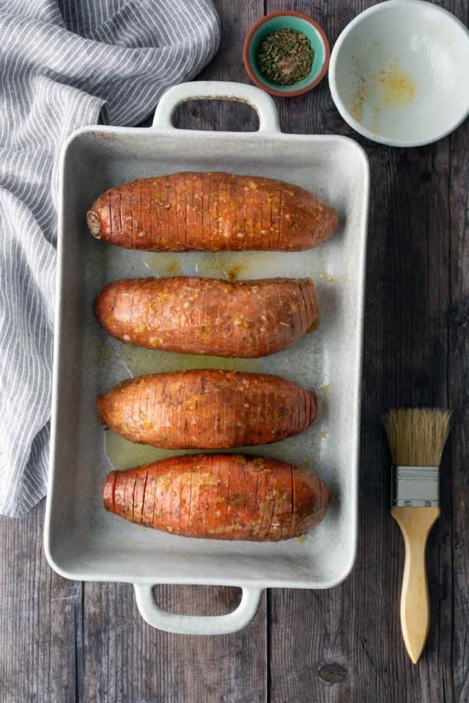 Four sliced sweet potatoes arranged in a white baking dish, with a small bowl of herbs, an empty bowl, and a basting brush nearby on a wooden surface.