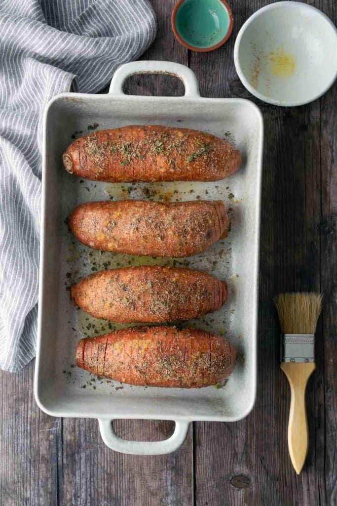 Four seasoned Hasselback sweet potatoes in a white baking dish, placed on a wooden table with a striped towel, empty bowls, and a basting brush nearby.