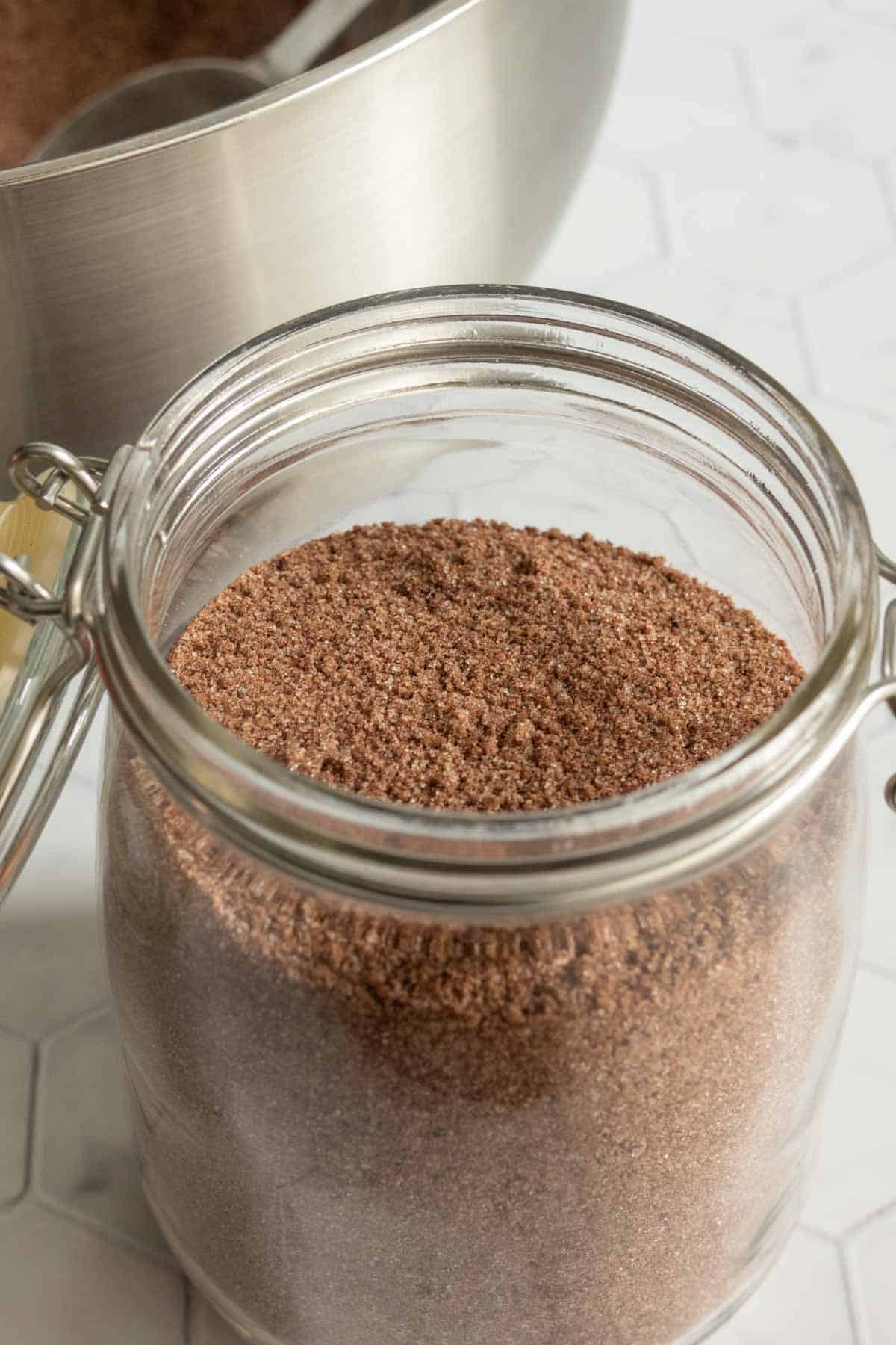A glass jar filled with cocoa powder mix on a white countertop, with a metal mixing bowl in the background.