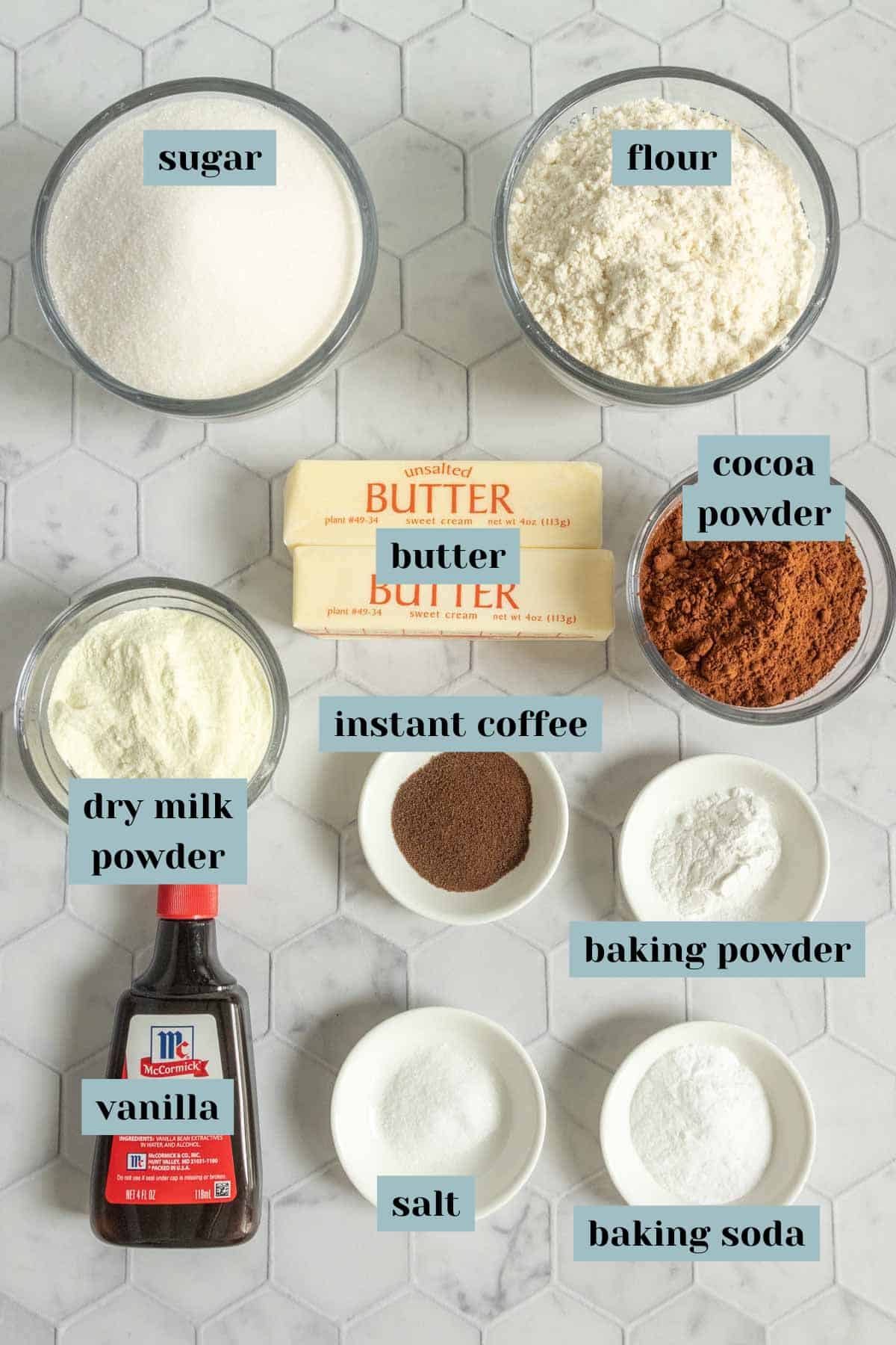 Various baking ingredients labeled in bowls and containers on a white hexagonal tile surface, including sugar, flour, cocoa powder, butter, dry milk powder, instant coffee, vanilla, salt, baking powder, and baking soda.