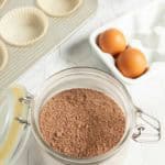 A jar of brown baking mix sits on a kitchen counter next to two eggs in a carton, a muffin tin with paper liners, and an electric mixer.