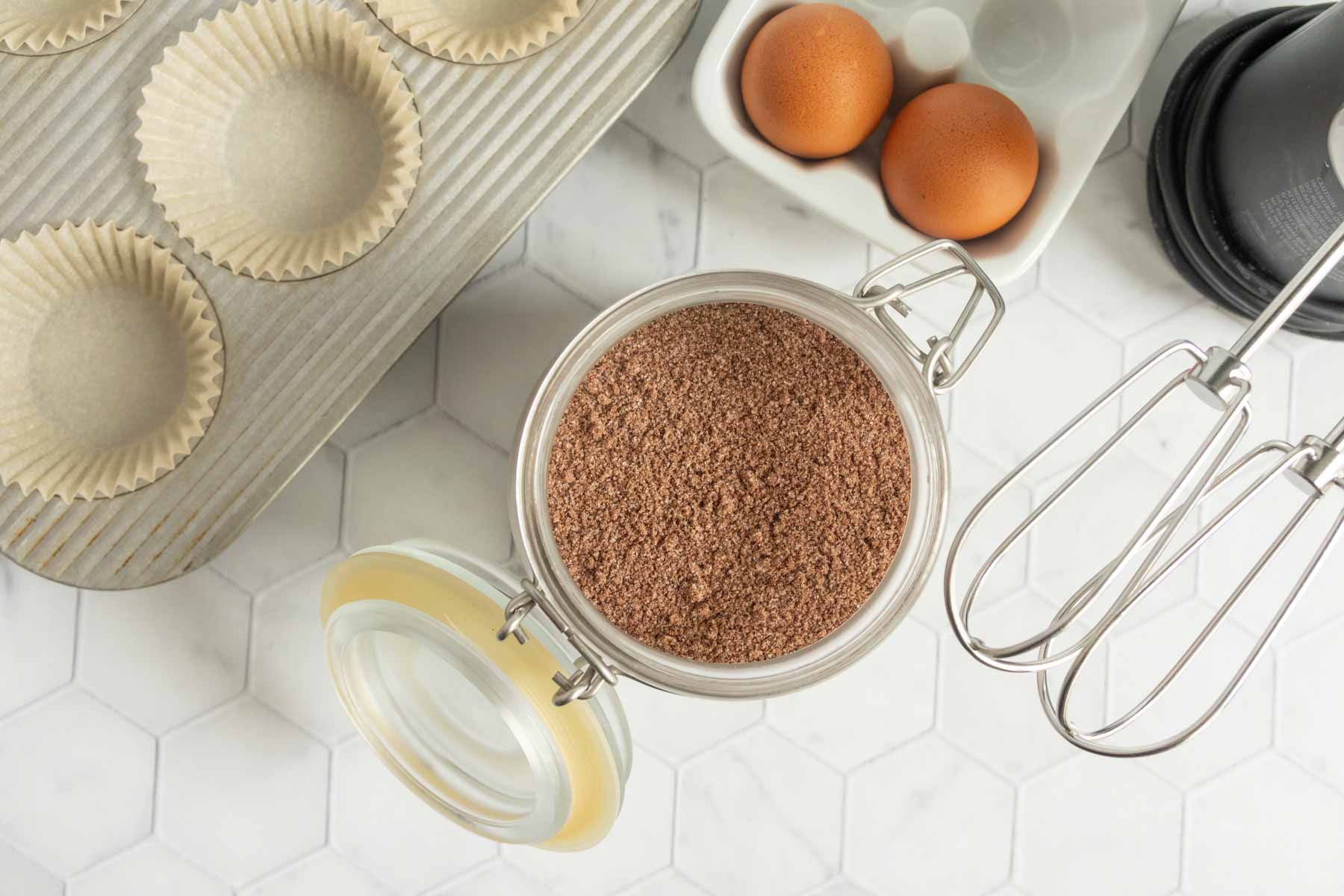 A jar of cocoa powder, two eggs in a dish, an electric mixer, cupcake liners in a tin, and a hexagon-tiled surface.