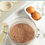 A glass jar filled with homemade devilโs food cake mix sits on a white counter next to three eggs and cupcake liners.