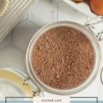 A glass jar of homemade devilโs food cake mix sits on a white countertop beside eggs and a muffin tray with paper liners.