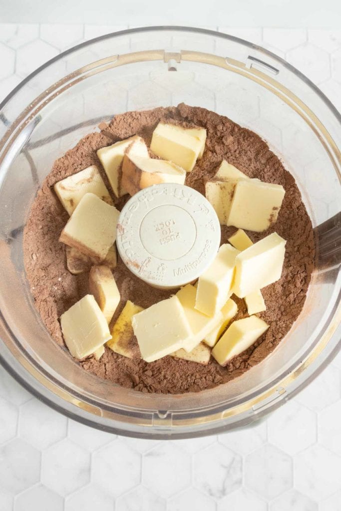 Cubed butter and cocoa powder in a food processor bowl, ready to be mixed, on a white hexagonal tiled surface.