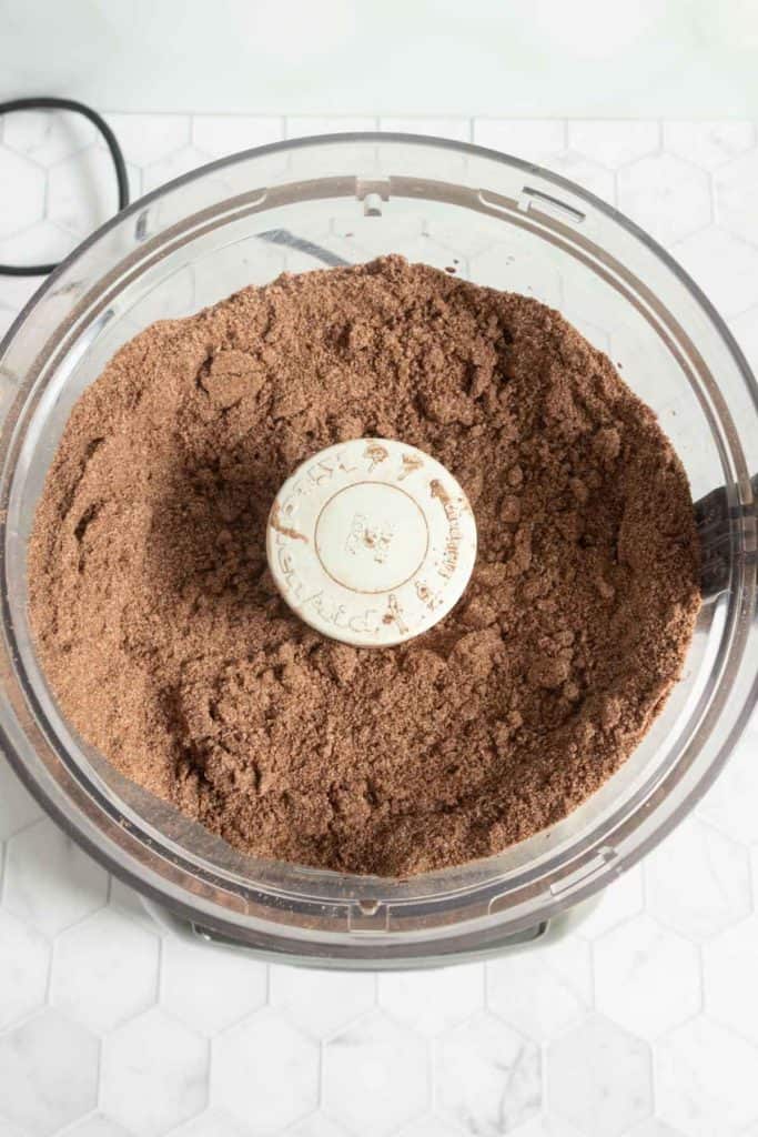 A food processor filled with a crumbly mixture of brown powder sits on a white hexagon-tiled countertop.