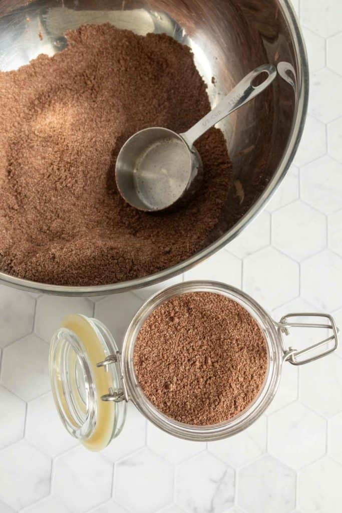 A metal bowl with a measuring cup and ground brown powder next to a glass jar filled with the same powder on a white hexagonal tile surface.