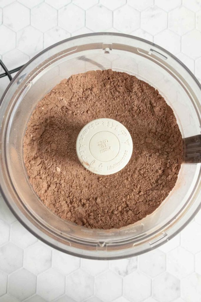 A food processor bowl filled with blended cocoa powder and dry ingredients sits on a white hexagonal tile countertop.