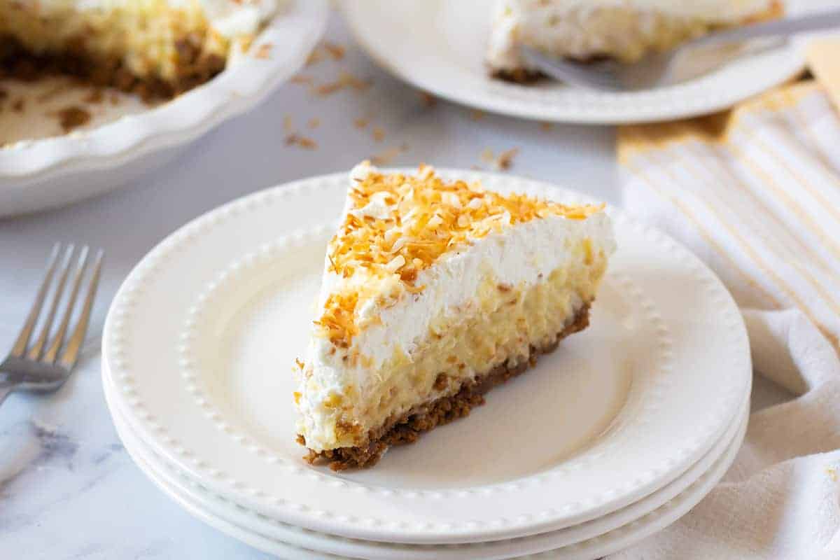 A slice of cream pie with a graham cracker crust and toasted coconut topping on a stack of white plates.