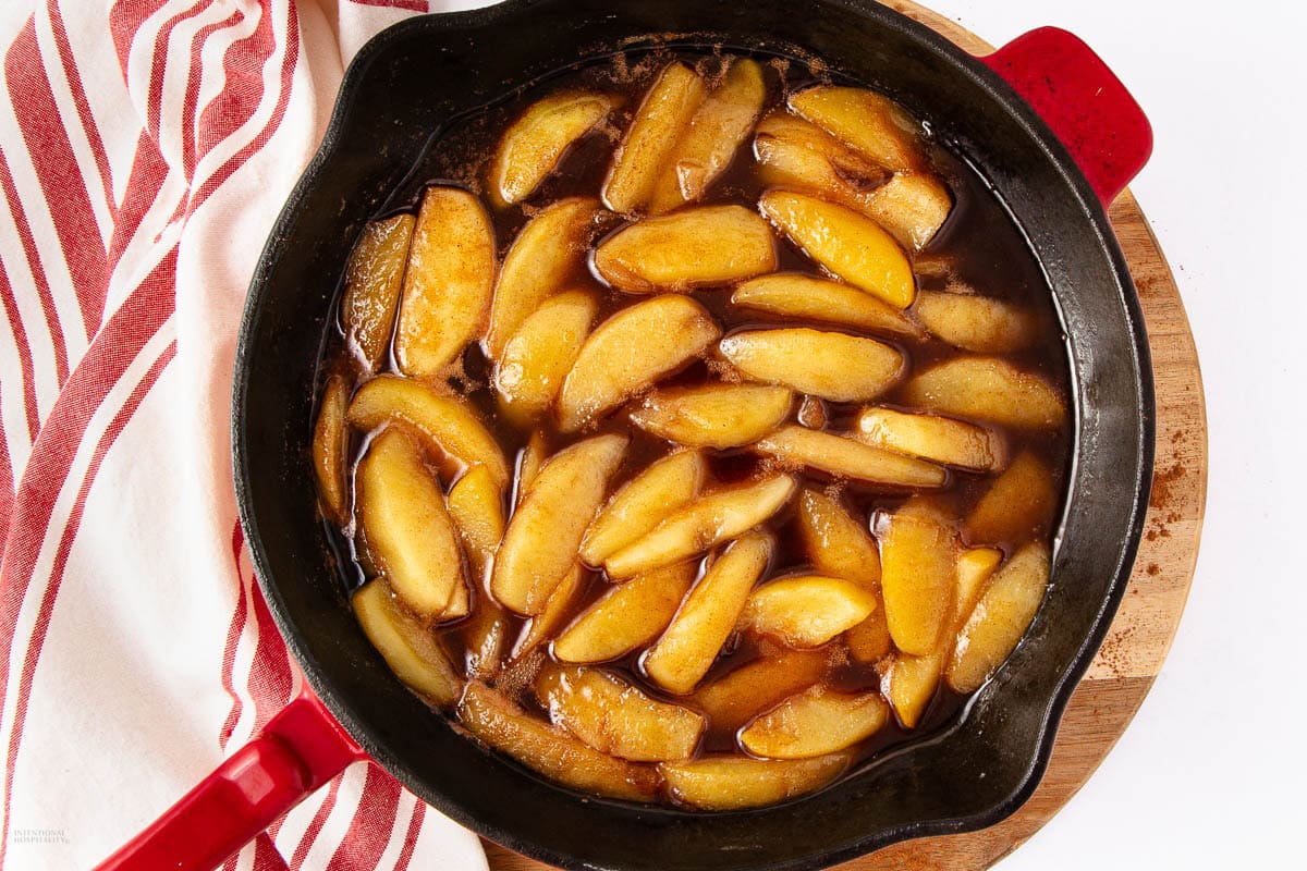 A cast iron skillet filled with cooked apple slices in a brown sauce sits on a wooden board next to a red and white striped towel.
