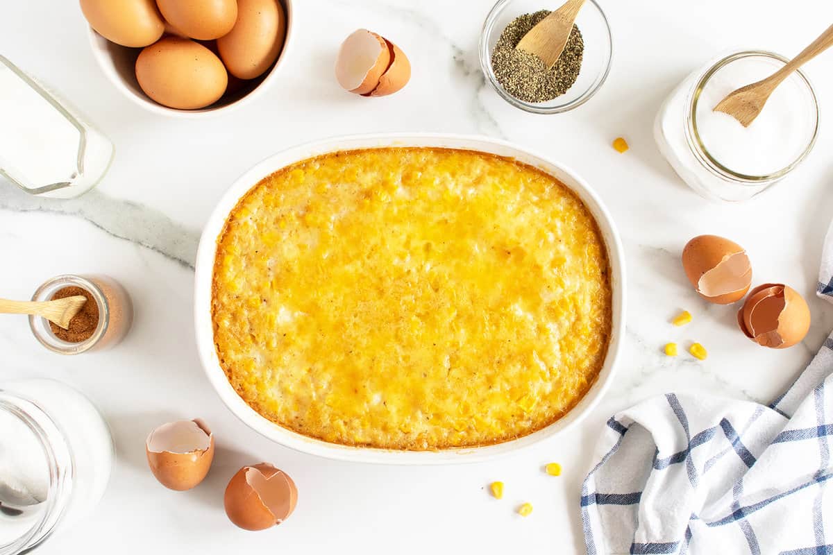 A baked casserole dish filled with cheesy cornbread sits on a marble countertop, surrounded by eggs, seasoning jars, and a striped kitchen towel.