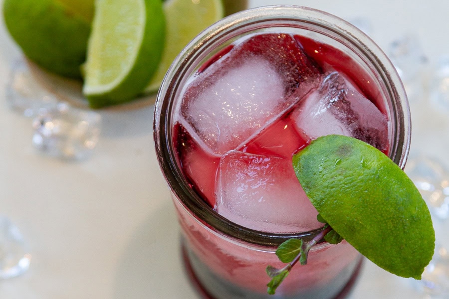 A glass filled with a red drink, ice cubes, and garnished with a lime wedge, with lime slices in the background.