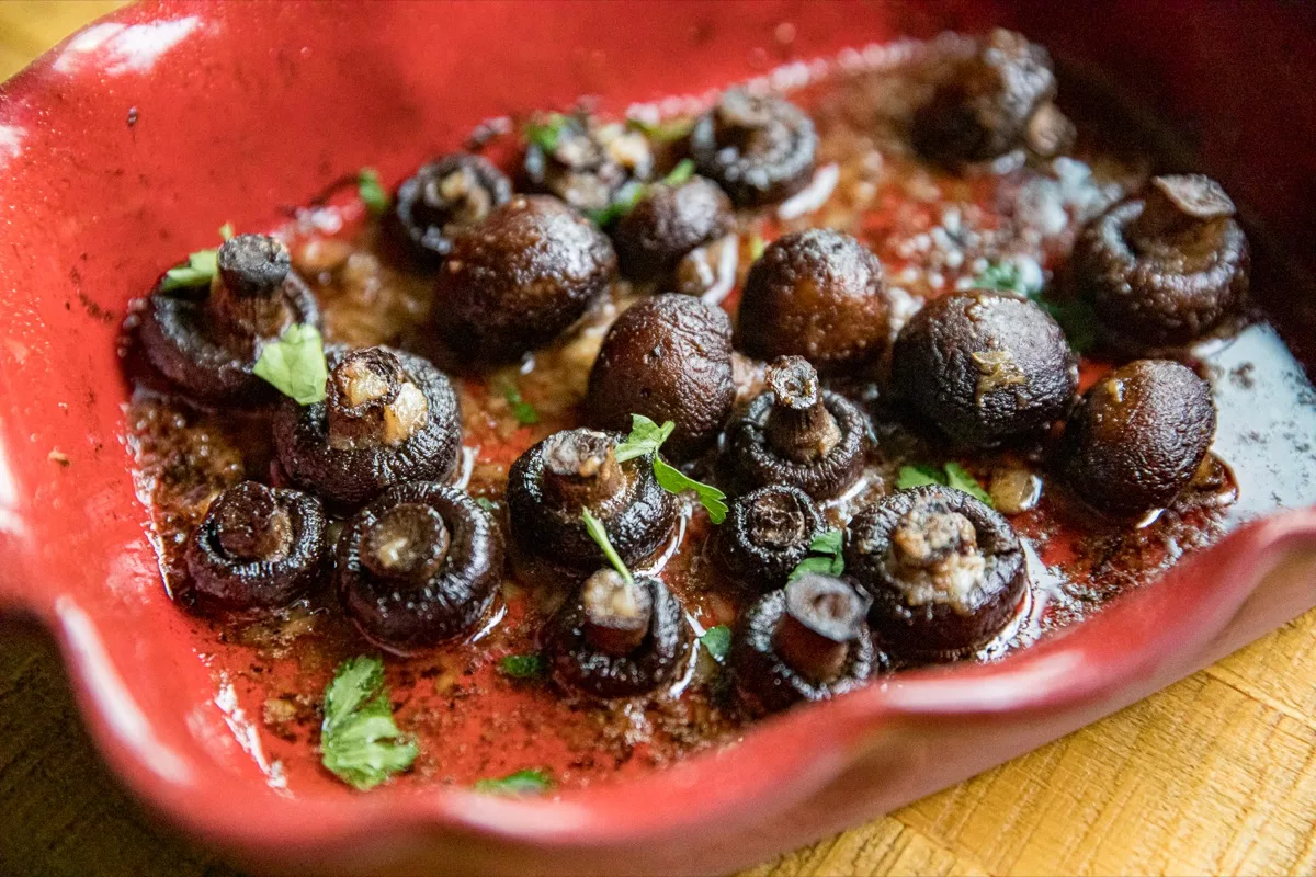 Roasted whole mushrooms garnished with chopped herbs sit in a red ceramic baking dish with visible juices on the bottom.