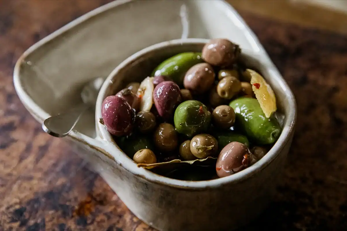 A ceramic bowl filled with assorted green and purple olives, garlic cloves, and herbs, placed on a rustic brown surface.