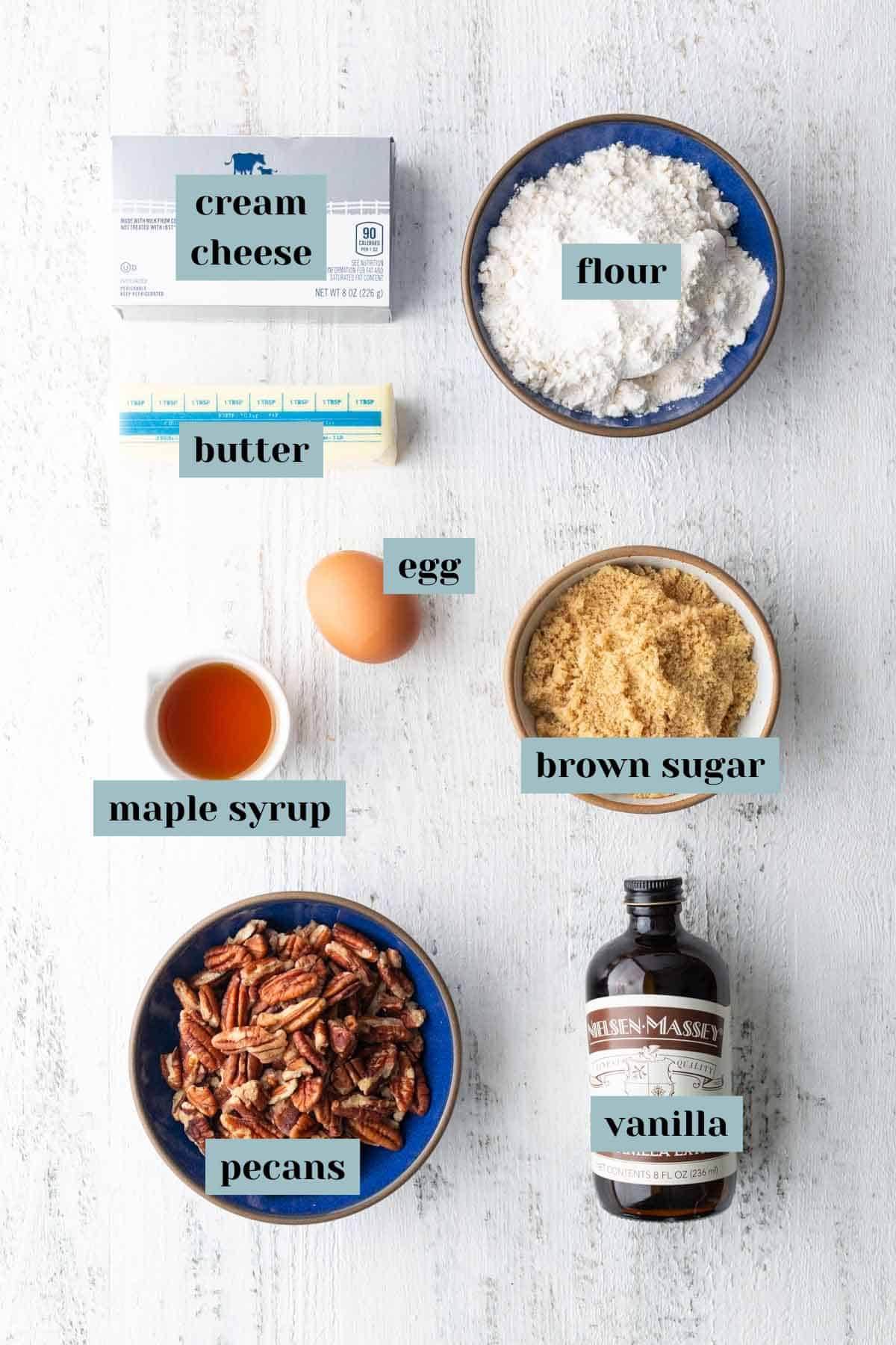 Top-down view of separated baking ingredients including cream cheese, flour, butter, egg, brown sugar, maple syrup, pecans, and a bottle of vanilla on a white surface.