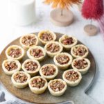A round board holds fifteen mini pecan tarts on a white table, with a glass of milk and decorative bottle brush trees in the background.
