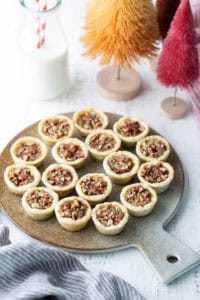 A round board holds fifteen mini pecan tarts on a white table, with a glass of milk and decorative bottle brush trees in the background.