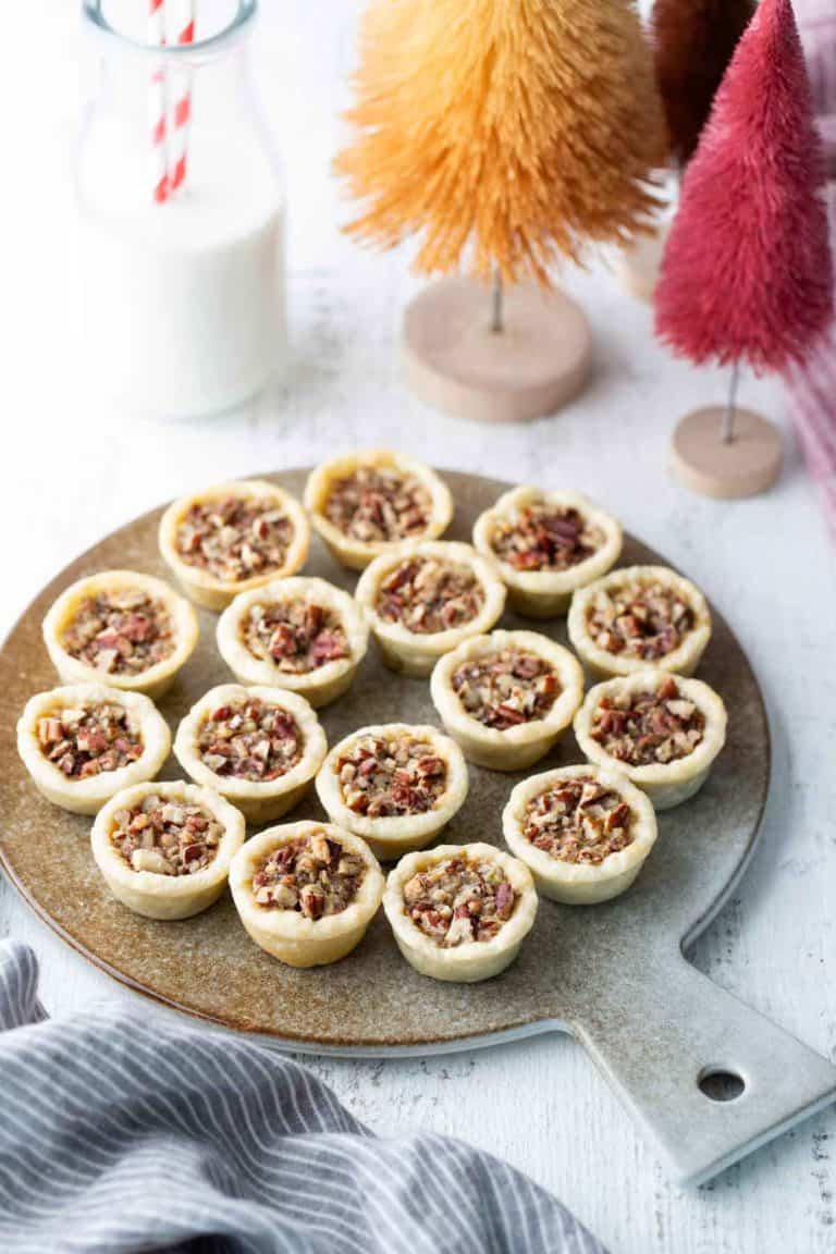 A round board holds fifteen mini pecan tarts on a white table, with a glass of milk and decorative bottle brush trees in the background.