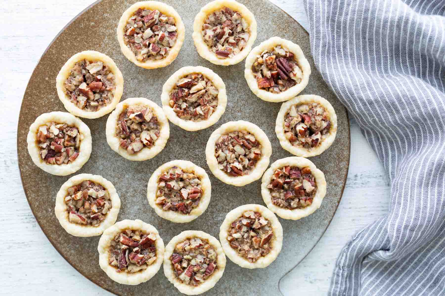 A round tray holds fifteen mini pecan tarts arranged in a circular pattern, next to a gray and white striped cloth on a white surface.