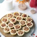 A round plate with fifteen pecan tassies, small tart-like desserts filled with chopped pecans, on a white table with candles and holiday decorations in the background.
