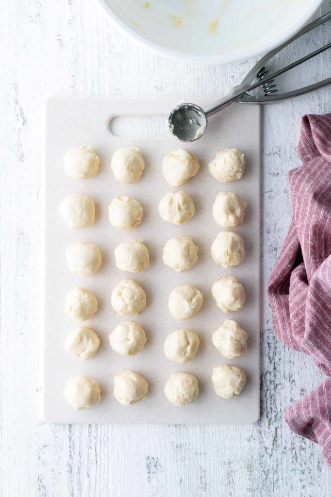 Twenty-four evenly spaced raw dough balls on a white cutting board with a metal scooper and a red striped cloth nearby.