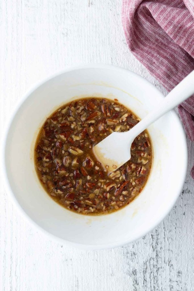 White mixing bowl with a spoon, containing a mixture of chopped pecans and a brown liquid, on a white wooden surface next to a red-striped cloth.