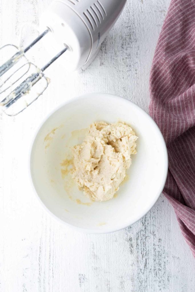A white bowl with cookie dough and a handheld electric mixer on a white surface next to a red striped cloth.