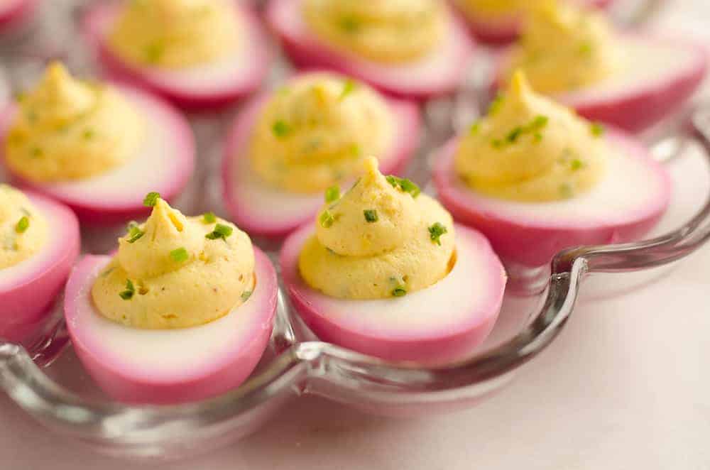 Halved deviled eggs with pink-stained whites, filled with a yellow yolk mixture and garnished with chopped green herbs, arranged on a glass serving tray.