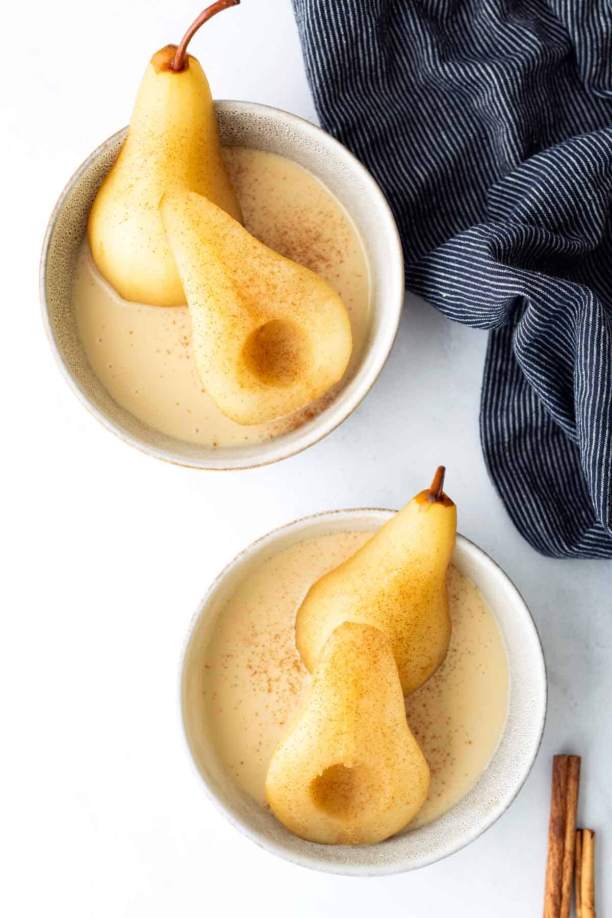 Two bowls with whole poached pears in a creamy sauce, sprinkled with cinnamon, next to a blue striped cloth and a cinnamon stick.
