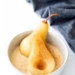 Two poached pear halves in a bowl with creamy sauce, dusted with cinnamon, with a striped cloth in the background.