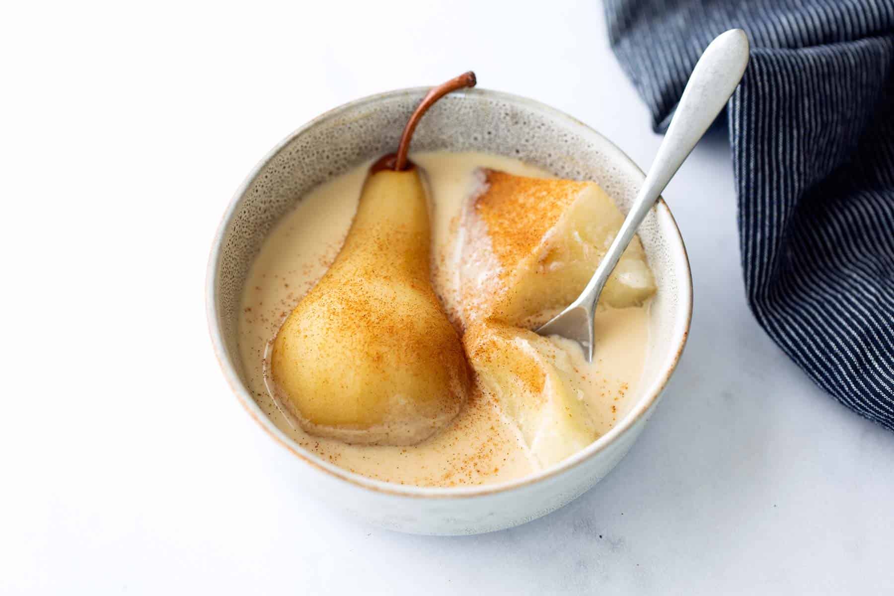 A bowl with a whole poached pear and a sliced piece in creamy sauce, sprinkled with cinnamon, with a spoon and a blue cloth napkin nearby.