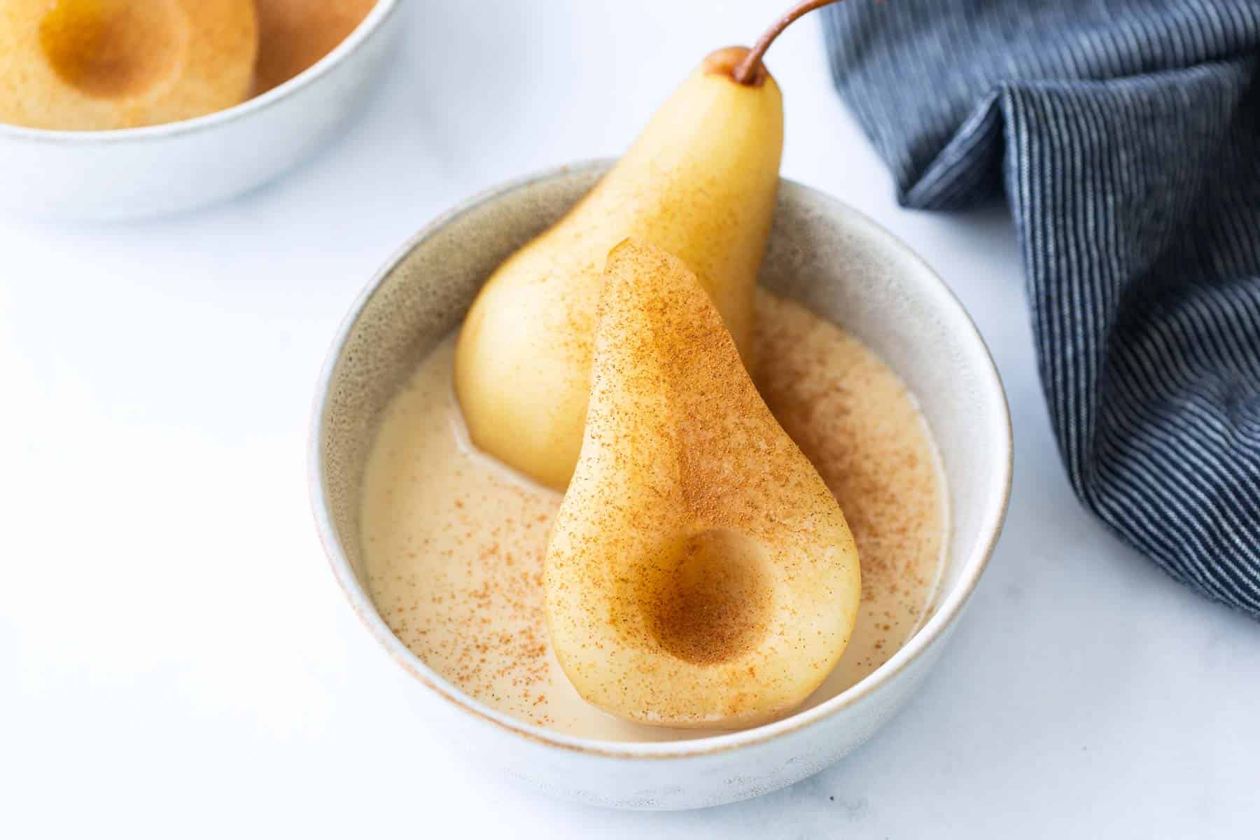 A bowl with two poached pear halves in creamy sauce, sprinkled with cinnamon, next to a folded striped cloth.