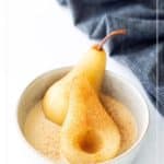 A bowl with two tea-poached pear halves in light sauce, sprinkled with cinnamon, placed on a white surface next to a dark striped cloth.