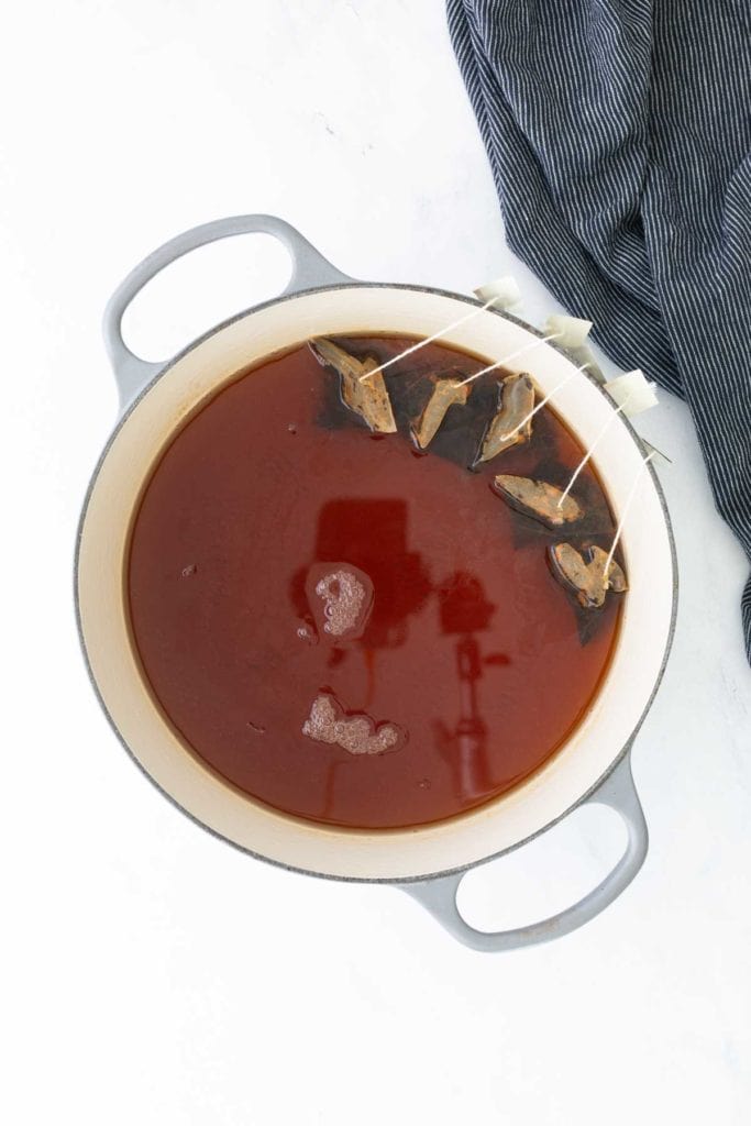 A large pot with tomato soup and four tea bags steeping on one side, placed on a white surface next to a dark striped cloth.
