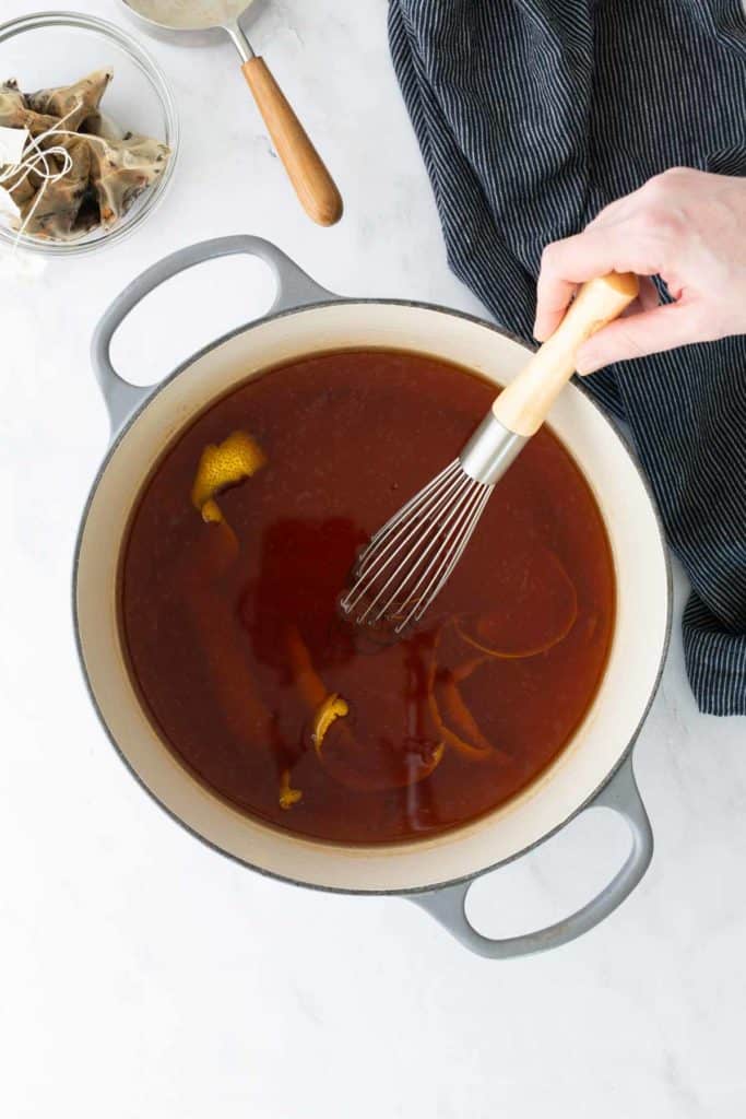 A hand uses a whisk to stir reddish-brown liquid with citrus peels in a large gray pot on a white countertop.