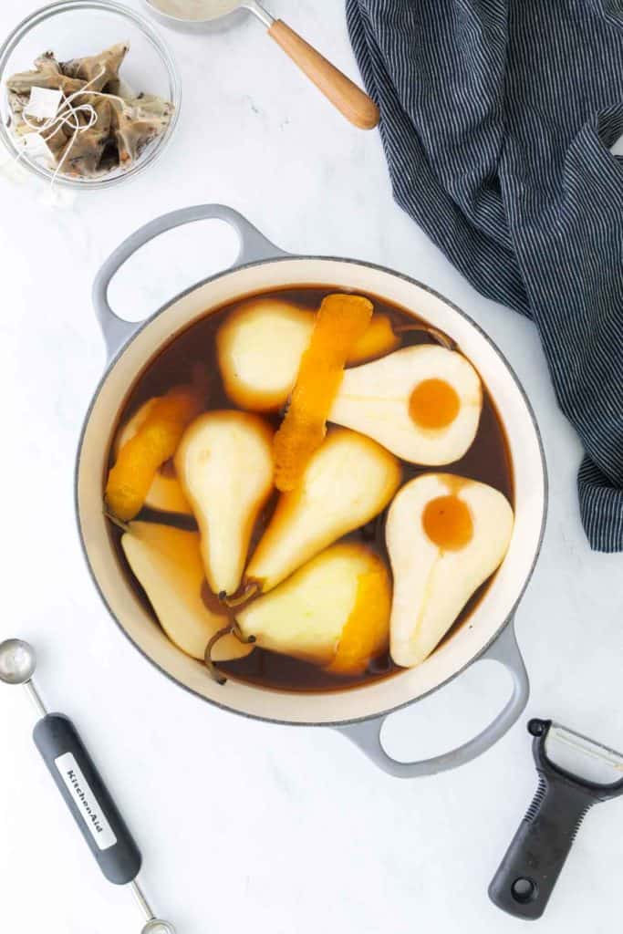A pot with halved pears, orange peels, and dark liquid sits on a white surface near a peeler, measuring spoon, striped cloth, and a bowl of spices.