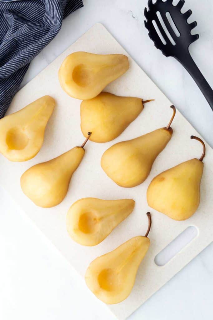 Eight peeled and halved pears with cores removed are arranged on a white cutting board next to a black slotted spoon and a dark striped cloth.